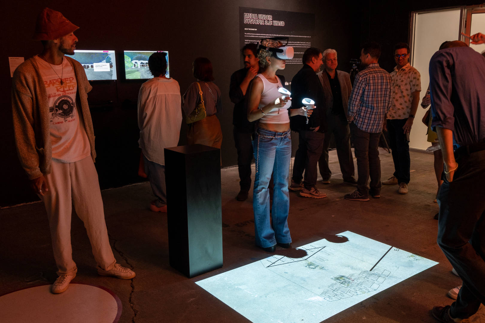 A woman using an Oculus standing over a projection of a metaverse scene, with people around at the MUD 3.0 exhibition