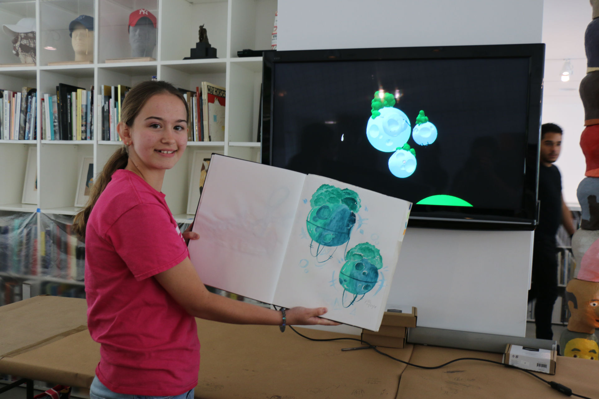A girl showing a drawing in a notebook in front of the 3D version of the same drawing in a screen.